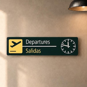 Könnte beinhalten: Ein rechteckiges Wandschild mit einem gelben Quadrat und einem schwarzen Flugzeugsymbol auf der linken Seite, dem Text "Departures" und "Salidas" in Weiß und Gelb sowie einer runden analogen Uhr auf der rechten Seite vor dunkelgrünem Hintergrund.