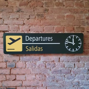 May include: A rectangular sign with a dark green background and white text that reads "Departures" and "Salidas" in yellow. A yellow square with a plane icon is on the left, and a clock is on the right. The sign is mounted on a brick wall.