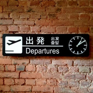 May include: Black rectangular sign with white text and symbols for Departures. The sign includes a stylized airplane icon, Japanese characters, and a clock face. The sign is mounted on a red brick wall.