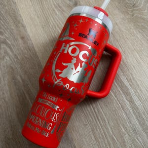 May include: A bright red Stanley tumbler with a handle and a clear straw. The cup features silver laser-etched graphics including the text Hocus Pocus, silhouettes of three figures, and the phrase Oh Look! Another Glorious Morning Makes Me Sick.