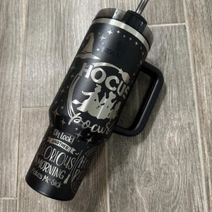 May include: Black stainless steel tumbler with a Hocus Pocus design. The tumbler has a black lid and a black handle. The design features the Sanderson sisters from the movie Hocus Pocus. The text on the tumbler reads "Hocus Pocus" and "Oh Look! Another Glorious Morning Makes Me Sick."