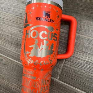 May include: Orange stainless steel travel mug with a black handle. The mug has a black and white graphic of the Sanderson sisters from the movie Hocus Pocus. The text "Hocus Pocus" is printed on the mug. The mug also has the Stanley logo and the text "I'm locked in another Hocus Pocus Morning, makes me sick."