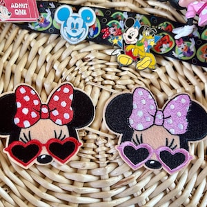 May include: Two Minnie Mouse patches with heart-shaped sunglasses. One patch has a red bow with white polka dots, the other has a lavender bow with white polka dots. Both have black ears and are on a woven background.
