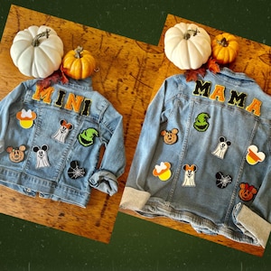 May include: Two light blue denim jackets decorated with embroidered patches. One jacket features the word MINI, the other MAMA. Both include patches of ghosts, pumpkins, spiderwebs, and candy corn, all styled with iconic mouse-ear silhouettes.