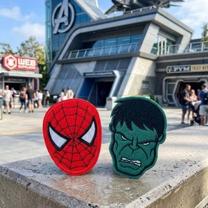 May include: Two embroidered patches featuring Marvel superheroes. One patch is red with a white spiderweb design and white eyes, resembling Spider-Man. The other is green, depicting the Hulk's face. The patches are displayed outdoors.