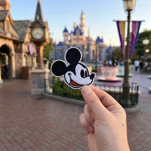May include: An embroidered fabric patch featuring the head of the cartoon character Mickey Mouse, depicted with black ears, a white face, and a red mouth, held up against a blurred background of a castle and park architecture.