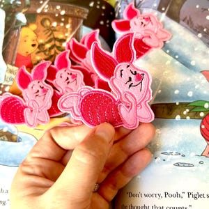 May include: A set of five pink embroidered iron-on patches featuring Piglet from Winnie the Pooh. The patches are in various poses, including Piglet thinking and Piglet smiling. The patches are on a book with a snowy background.