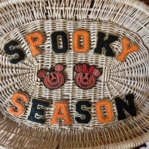 May include: A set of embroidered patches featuring the text SPOOKY SEASON in alternating black and orange varsity-style letters with gold glitter borders, alongside two pumpkin-shaped patches with mouse ears, one with a bow.