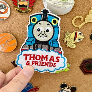 May include: A blue, white, and black Thomas the Tank Engine patch with the text "THOMAS & FRIENDS" in red. The patch is surrounded by other character pins, including a Baby Yoda pin.