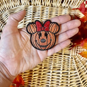 May include: An embroidered patch shaped like a pumpkin with mouse ears and a red bow. The pumpkin is orange with black stitching details, including a nose, eyes, and a mouth. The entire patch is outlined in black thread against a dark background.