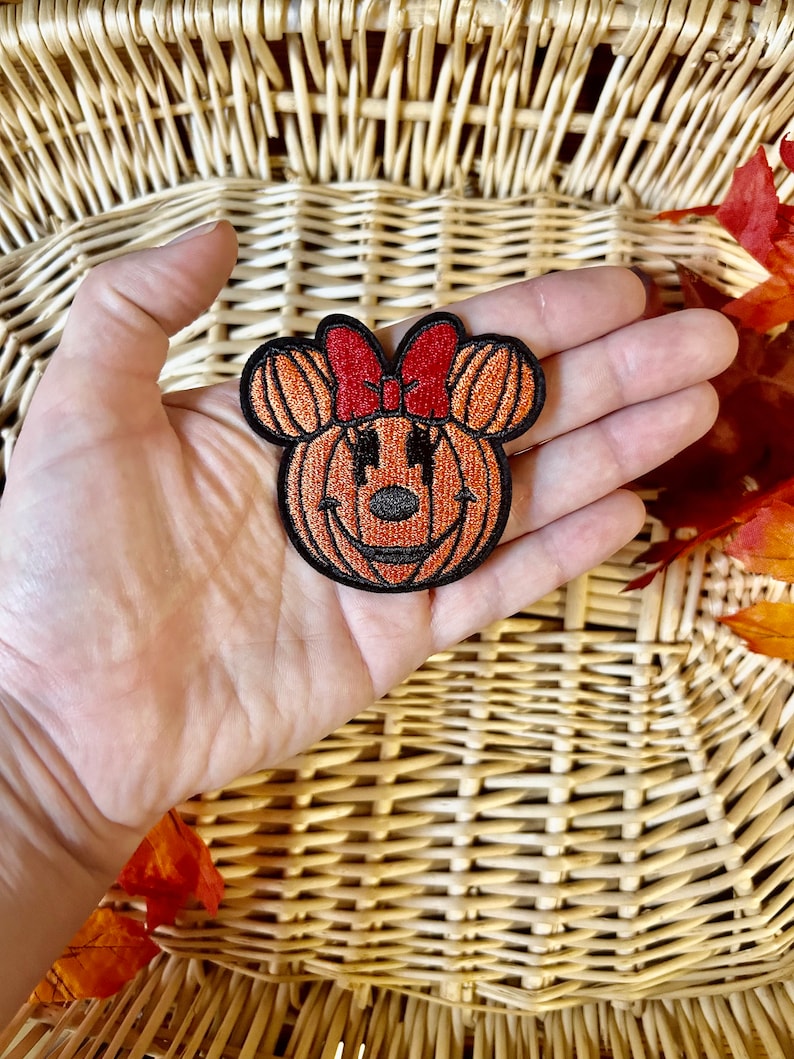 May include: An embroidered patch shaped like a pumpkin with mouse ears and a red bow. The pumpkin is orange with black stitching details, including a nose, eyes, and a mouth. The entire patch is outlined in black thread against a dark background.