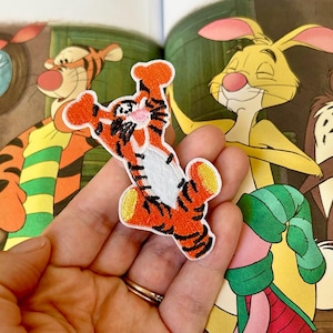 May include: Embroidered patch featuring Tigger from Winnie the Pooh. The patch is orange with black stripes, a white belly, and yellow feet. The patch is held in front of a book with illustrations of other characters.