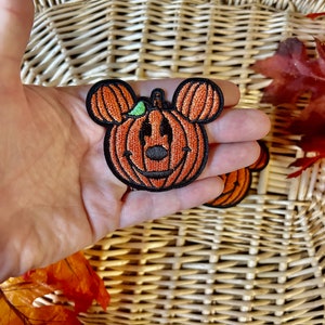 May include: An embroidered patch shaped like a pumpkin with round ears, featuring orange thread for the pumpkin segments, a black stitched outline, a small green leaf detail, and a black nose and mouth design resembling a classic character silhouette.