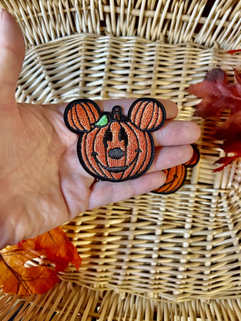 May include: An embroidered patch shaped like a pumpkin with round ears, featuring orange thread for the pumpkin segments, a black stitched outline, a small green leaf detail, and a black nose and mouth design resembling a classic character silhouette.