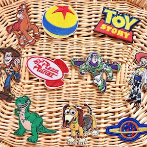 May include: A collection of embroidered patches featuring characters and logos from the animated film "Toy Story." Patches include Woody, Buzz Lightyear, Jessie, Rex, and the Pizza Planet logo. The patches are on a woven wicker surface.