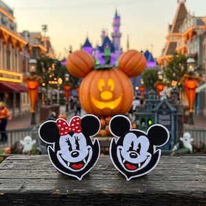 May include: Two embroidered patches featuring the faces of a black and white mouse character with vampire fangs. One patch includes a red bow with white polka dots. The designs are circular with pointed bottoms, showcasing detailed stitching.