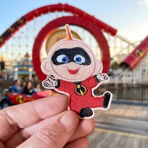 May include: Embroidered patch featuring Jack-Jack Parr from The Incredibles. The patch depicts Jack-Jack in a red suit with a black mask, blue eyes, and a smiling expression. The background shows a roller coaster.