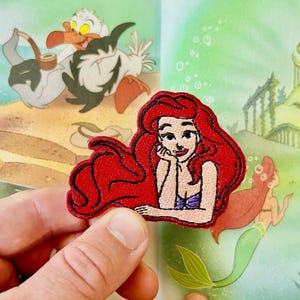 May include: A red-haired mermaid with a seashell bra and a crown, sitting with her hand on her chin. The mermaid is embroidered on a fabric patch.