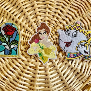 May include: Three embroidered patches featuring characters from the Disney movie Beauty and the Beast. The patches are on a woven wicker background. The patches are of Belle, the enchanted rose, and Mrs. Potts.