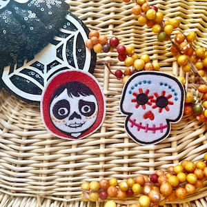 May include: A black sequined Minnie Mouse ear headband with a white spiderweb design, and two embroidered patches. One patch features a character from the movie Coco, and the other is a sugar skull. The items are arranged on a woven basket.
