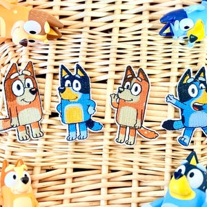 May include: A collection of Bluey and Bingo character patches and figurines. The patches feature embroidered designs of the cartoon dogs in various poses, with orange, blue, and black colors. The figurines are also in the likeness of the cartoon characters.