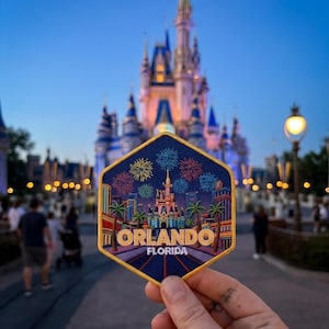 May include: A navy blue, hexagonal patch featuring a colorful illustration of Orlando, Florida, with fireworks, palm trees, and a castle. The word "ORLANDO" is in gold lettering, with "FLORIDA" below. The patch is held in front of a blurred background of a castle and a blue sky.