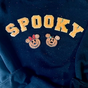 May include: A black sweatshirt featuring the word SPOOKY in orange glittery varsity-style letters. Below the text are two embroidered pumpkin patches shaped like mouse heads, one with a red bow and one with a small green leaf accent.