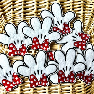 May include: White felt Mickey Mouse hand cutouts with black outlines and red polka dot bows. These craft supplies are perfect for DIY projects, party decorations, or themed crafts. The hands are arranged on a woven basket.