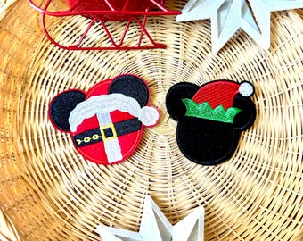 May include: Two embroidered patches featuring circular silhouettes with mouse ears. One patch depicts a red Santa suit with a white trim and black belt. The second patch shows a black silhouette wearing a red and green elf hat with a white pom-pom.