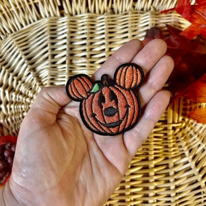 May include: An embroidered patch featuring an orange pumpkin with a black outline and black stitched facial features, including a nose and a wide smile. The pumpkin has two smaller orange pumpkin-shaped ears on top and a small green leaf accent.