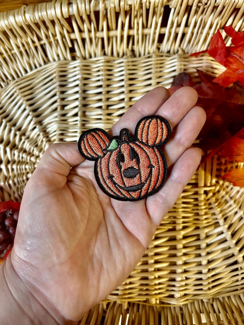 May include: An embroidered patch featuring an orange pumpkin with a black outline and black stitched facial features, including a nose and a wide smile. The pumpkin has two smaller orange pumpkin-shaped ears on top and a small green leaf accent.