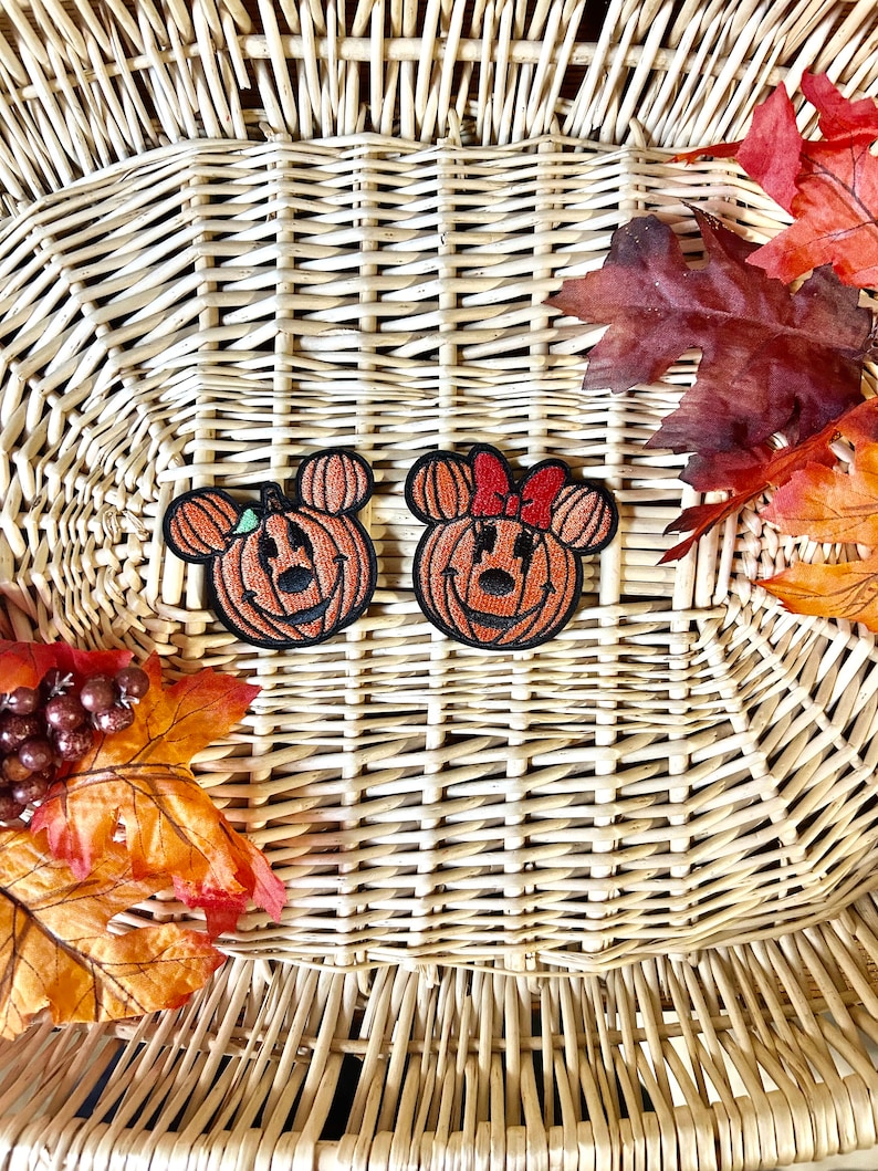 May include: Two embroidered patches featuring orange pumpkin shapes with black outlines. One patch has mouse ears and a small green leaf, while the other has mouse ears and a red bow. Both patches display a carved jack-o'-lantern face design.