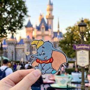 May include: A Dumbo patch, featuring the cartoon elephant with a yellow hat, red collar, and large pink ears. The patch is held in front of a castle and theme park setting.