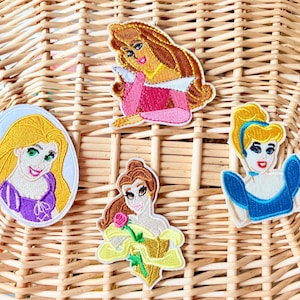 May include: Four embroidered iron-on patches featuring Disney princesses. The patches are on a woven wicker background. The princesses are Rapunzel with long blonde hair, Belle with brown hair, Cinderella with blonde hair, and Sleeping Beauty with brown hair.