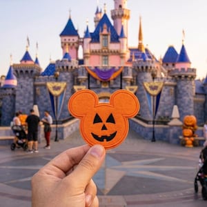 May include: An orange felt patch shaped like a Mickey Mouse head with a black jack-o'-lantern face featuring triangular eyes and a jagged mouth. The patch is held up against a backdrop of a large castle with blue turrets and pink walls.