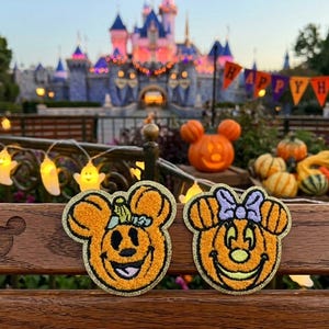 May include: Two embroidered patches featuring orange pumpkin-shaped characters with mouse ears. The left patch shows a pumpkin with a green stem, while the right patch features a pumpkin with a purple bow. Both have gold glitter borders.