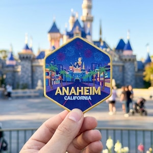 May include: A hexagonal embroidered patch featuring a nighttime scene with a castle, palm trees, and fireworks. The text ANAHEIM is stitched in large yellow letters, with CALIFORNIA in smaller white letters below, all bordered in gold thread.