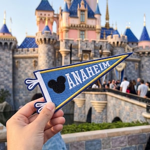 May include: A blue felt pennant flag with a white border and yellow trim. The pennant features a black silhouette of a mouse head icon and the white text ANAHEIM printed in capital letters. The flag has two white fabric ties on the left side.