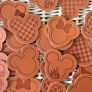May include: A collection of brown leather coasters with various designs, including Mickey Mouse ears, checkered patterns, and a castle silhouette.