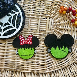 May include: Two embroidered patches shaped like mouse ears with green Frankenstein-style faces. One patch features a red and white polka dot bow, while the other has a stitched scar. Both have black ears and silver bolts on the sides.