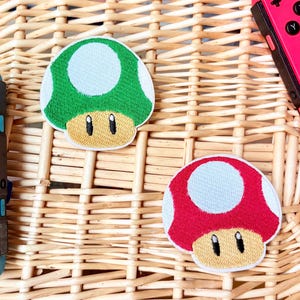 May include: Two embroidered mushroom patches, one green and one red, with white spots and tan faces. A teal and a pink game controller are also visible. The items are on a woven wicker surface. The patches are likely for video game enthusiasts.