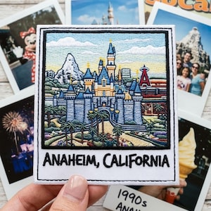 May include: A square embroidered patch featuring a detailed scene of a blue and yellow castle with a mountain and a red letter A logo in the background. The patch is framed in black and white with the text ANAHEIM, CALIFORNIA printed at the bottom.