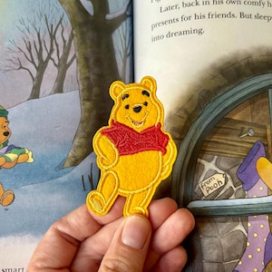 May include: A yellow Winnie the Pooh patch with a red shirt is held in front of a book illustration. The patch features embroidered details. The book illustration shows a winter scene with other Winnie the Pooh characters.