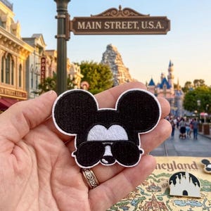 May include: A black and white embroidered patch featuring the iconic mouse head silhouette wearing dark sunglasses. The patch has a white border and is held up against a blurred background of a theme park street and castle.
