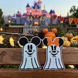 May include: Two embroidered patches featuring Mickey and Minnie Mouse as ghosts. Mickey has black ears and a smiling face, while Minnie has a bow. The patches are light blue with black outlines. The background includes a castle and Halloween decorations.