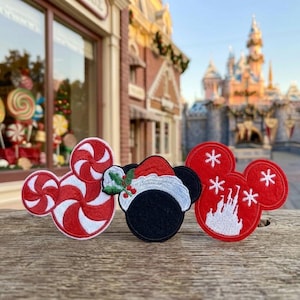 May include: Three embroidered patches shaped like Mickey Mouse ears. The left patch features a red and white peppermint swirl pattern. The center patch is black with a Santa hat and holly. The right patch is red with white snowflakes and a castle.