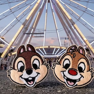 May include: Two embroidered patches featuring Chip and Dale, the Disney chipmunks, with a Ferris wheel in the background. The patches are brown, tan, and white, with smiling faces. The background includes a fairground with rides and people.