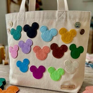 May include: A beige canvas tote bag featuring numerous colorful felt Mickey Mouse head appliques in shades of teal, black, light blue, yellow, pink, purple, coral, brown, green, white, dark blue, magenta, and lime green, with two small pins.