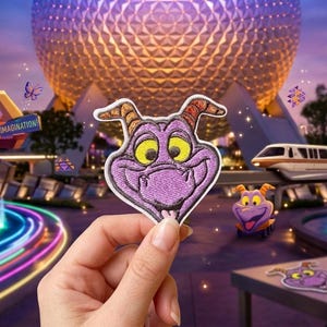 May include: A purple embroidered patch of a cartoon dragon character with yellow eyes and orange horns. The patch is held in front of a theme park with a large sphere and monorail in the background. Other patches are on a table.