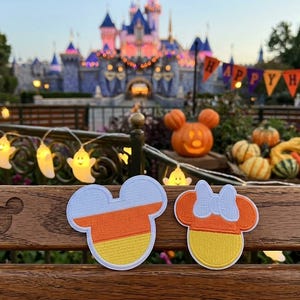 May include: Two embroidered patches shaped like mouse heads with candy corn color patterns of white, orange, and yellow. One patch features a simple silhouette, while the other includes a small bow detail at the top, both with white borders.